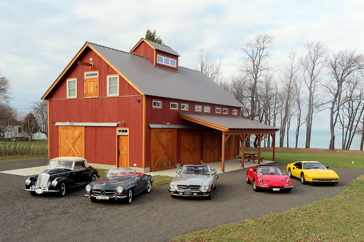 An exterior picture of the Lake Erie Auto Barn, designed and built by Geobarns, with barn red wood siding, gray metal roof, Douglas fir trim, cupola, and Lake Erie in the background.
