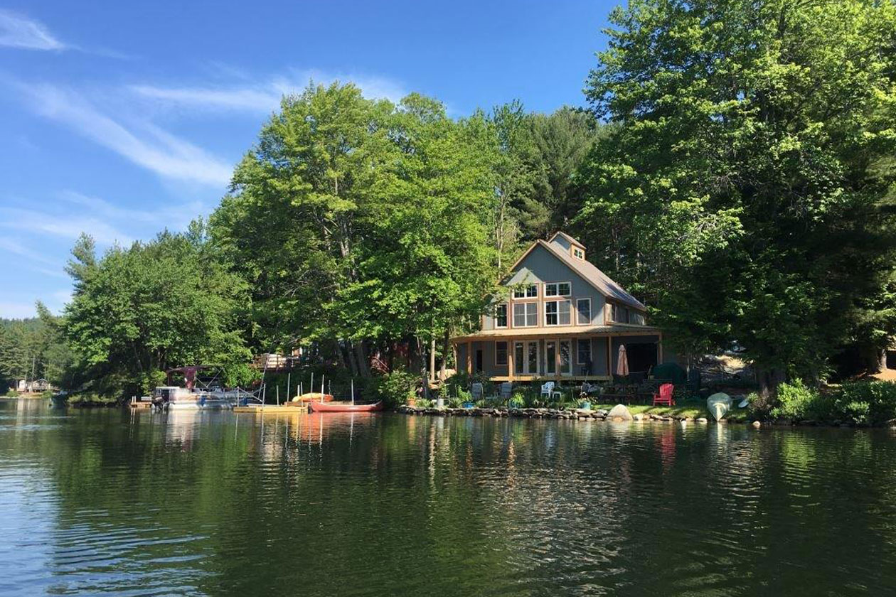 A picture of the Lake House and Barn, designed and built by Geobarns on a New Hampshire Lake, with gray wood siding, burnishied bronze metal roof, wide front porch, cupola, attached car barn, and boat dock.