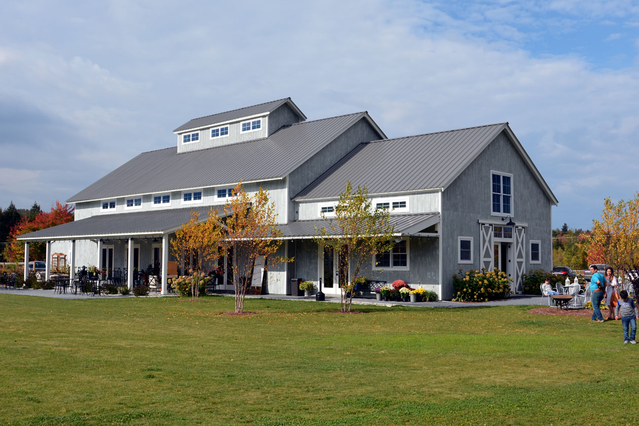 A picture of the Barn at Smuggler's Notch in Stowe, Vermont, a wedding and event barn designed and built by Geobarns, with gray stained wood siding, gray metal roof, cupola, rolling barn doors, event lawn, and Vermont mountains in the background.