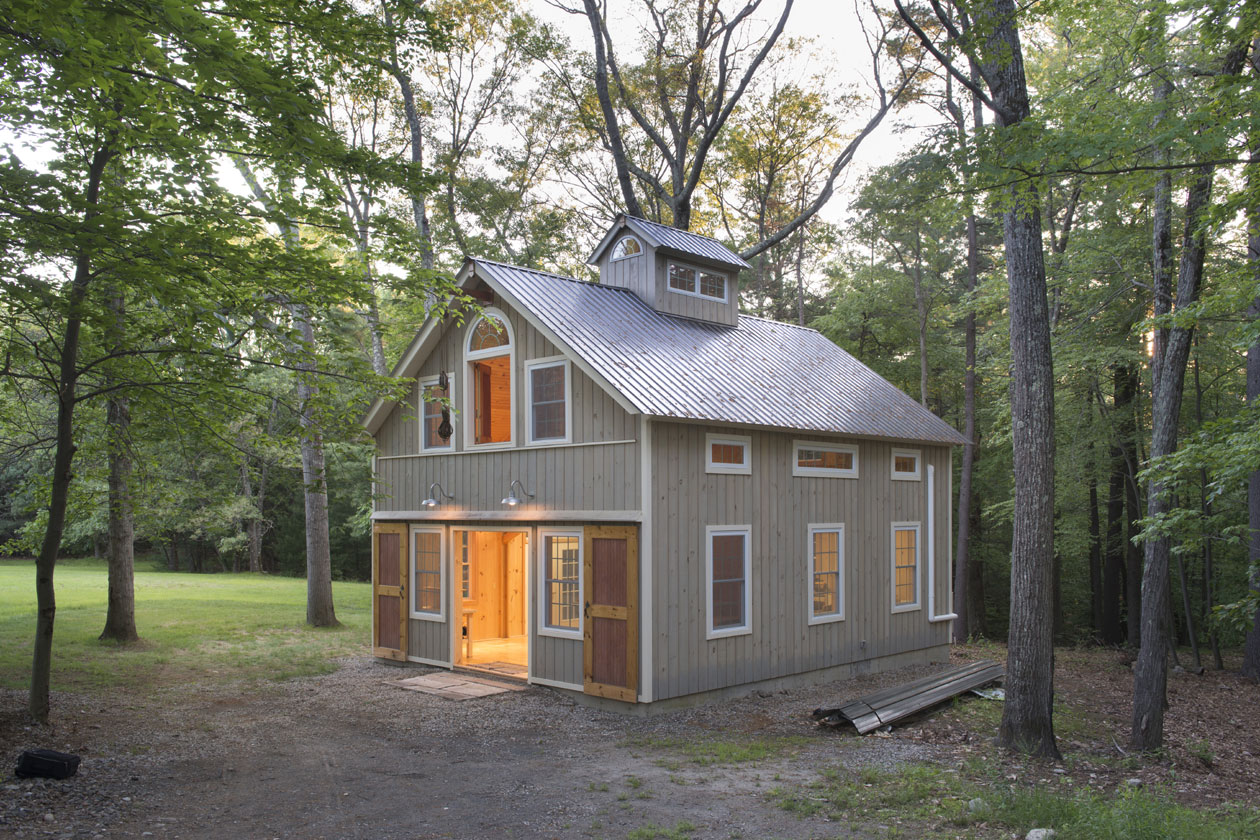 An exterior picture of the Woodworking Temple, design and built by Geobarns, with washed gray wood siding, silver metal roof, rolling barn doors, hayloft door, and cupola.