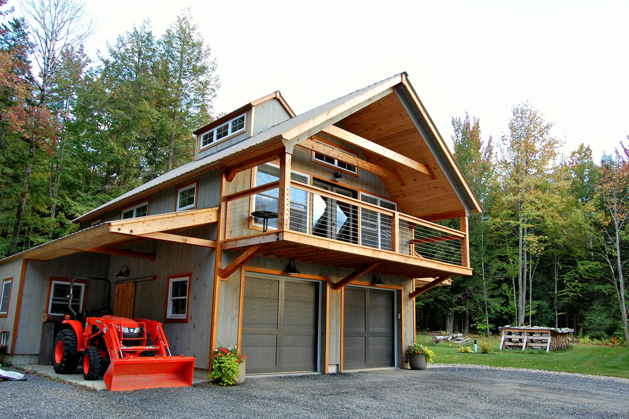An exterior picture of the Mountain Carriage House, designed and built by Geobarns, with gray wood siding, cantilevered front porch, and side awning to park a Kubota tractor.
