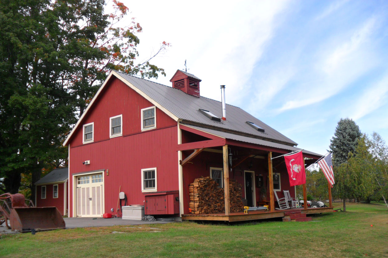 An exterior picture of the MLB Batmaker's Barn, designed and built by Geobarns, with barn red wood siding, gray metal roof, wide front porch, side garage, and cupola.