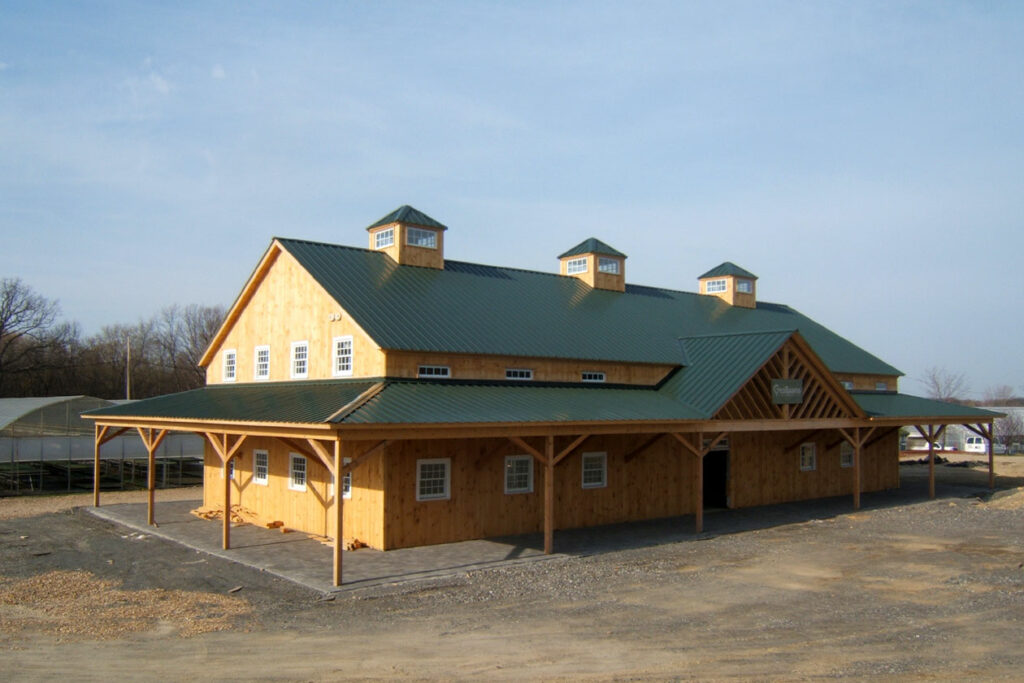 An exterior picture of the Green Thumb Nursery in Massachusetts, designed and built by Geobarns, with naturally weathered wood siding, green metal roof, three cupolas, and wrap-around porch.