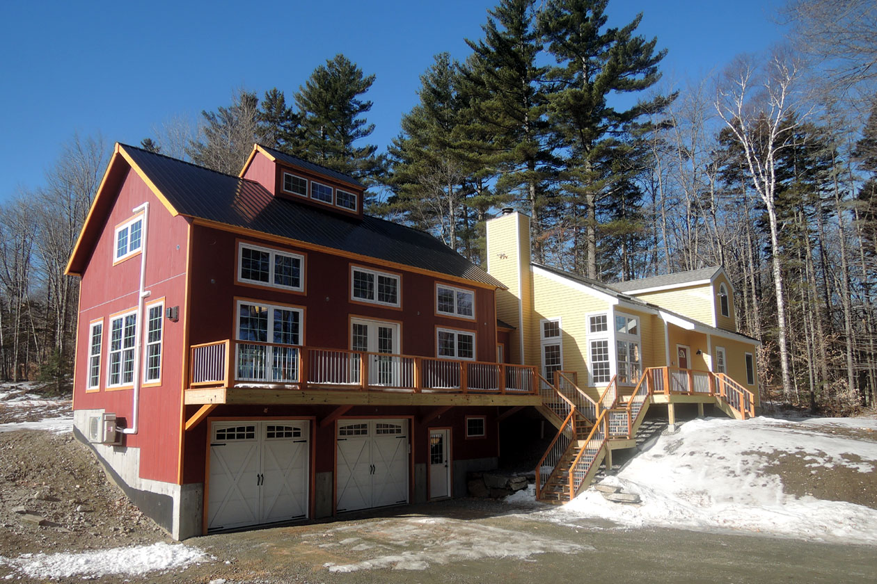 An exterior picture of an home office and entertainment addition, designed and built by Geobarns, with barn red wood siding, black metal roof, clerestroy windows, cupola, two-car garage, with a mudroom and a large deck.