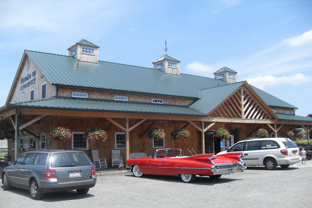 An exterior picture of the Green Thumb Nursery in Massachusetts, designed and built by Geobarns, with naturally weathered wood siding, green metal roof, three cupolas, and wrap-around porch.