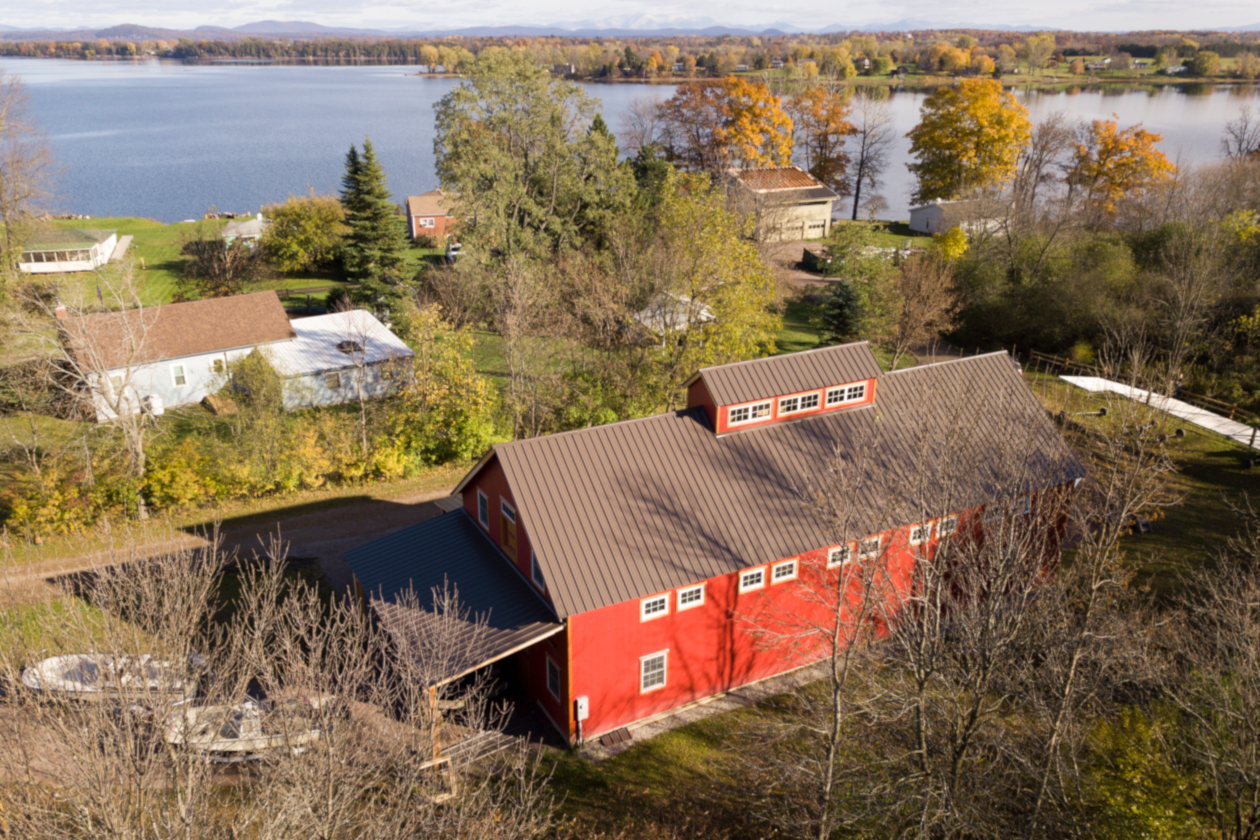 An aerial picture of the Lake Champlain Boat Barn, designed and built by Geobarns, with barn red woodsiding, gray metal roof, monitor cupola, with Lake Champlain in the background.