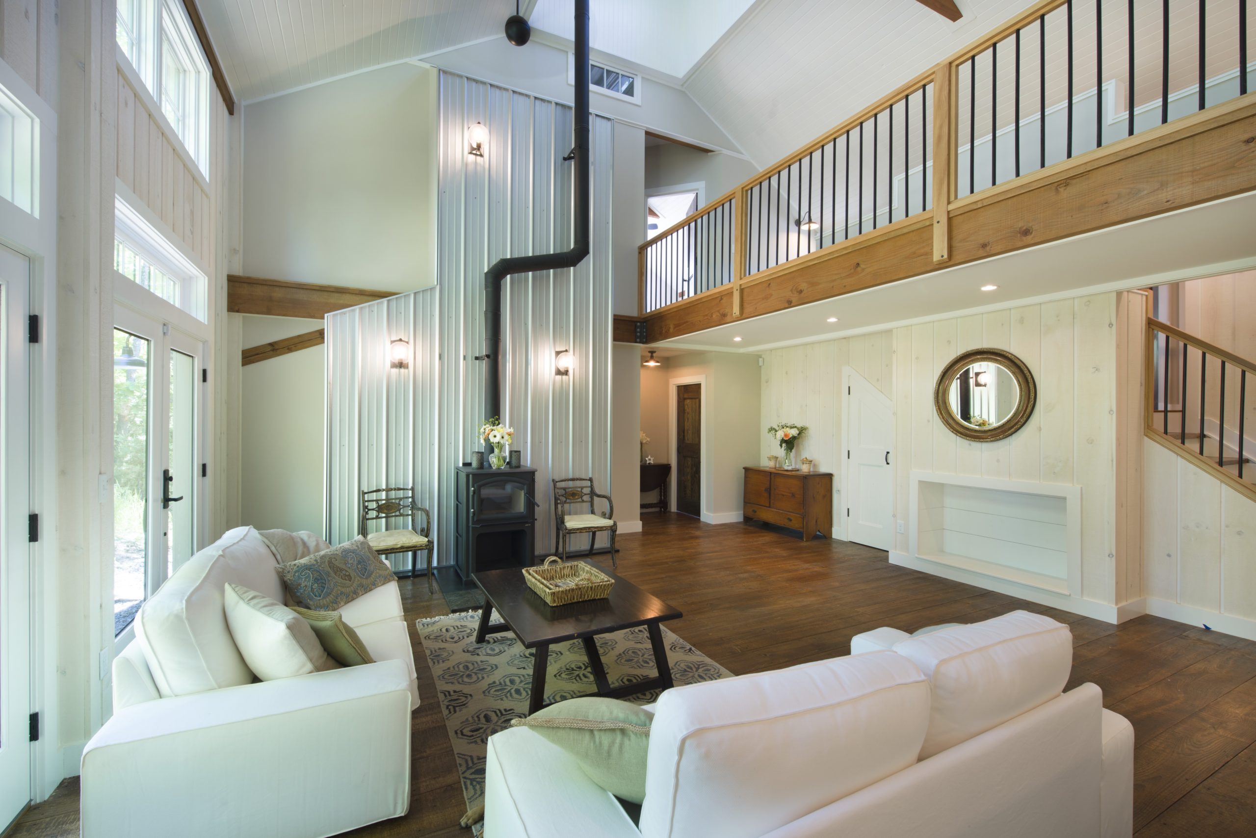 An interior picture of the living room of the Mountaintop Family Home, designed and built by Geobarns, with vaulted ceilings, exposed timber framing, metal firebreak behind a wood stove, mezzanine catwalk, and cupola high above.
