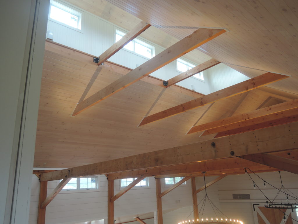 A picture of the ceiling of a Wedding Barn in Vermont showing the framing timbers, the wood ceiling, and sunlight streaming through the cupola