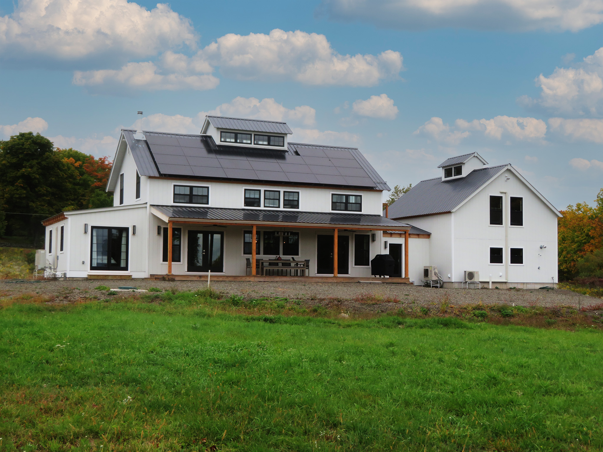 An exterior picture of the Catskills Modern Farmhouse, designed and built by Geobarns, with white wood siding, black metal roof, wide veranda, solar panels, and cupola.