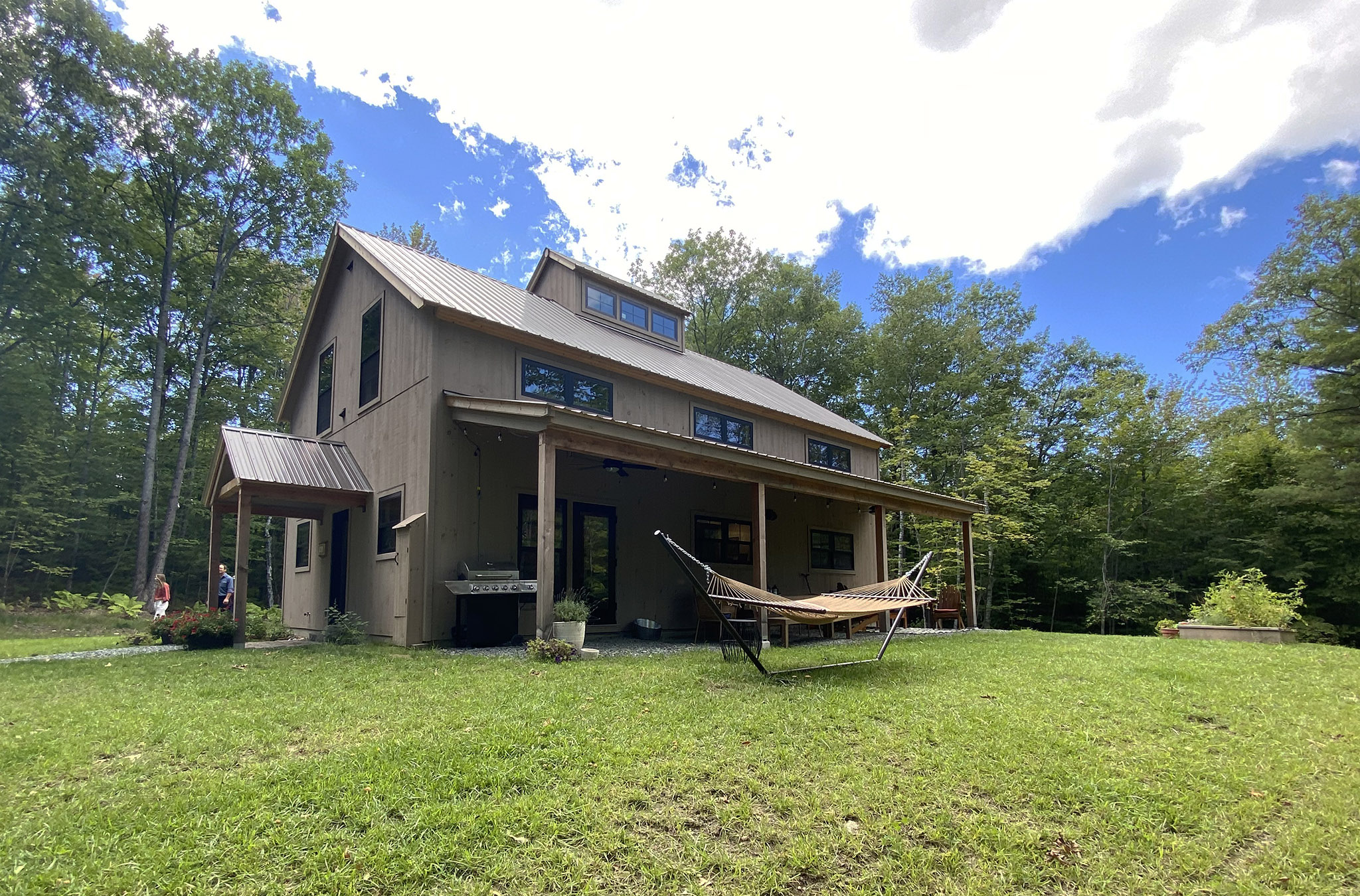 An exterior picture of the Forest Glade Retreat home, designed and built by Geobarns, with mocha wood siding, burnished bronze metal roof, front porch, side porch, and cupola.