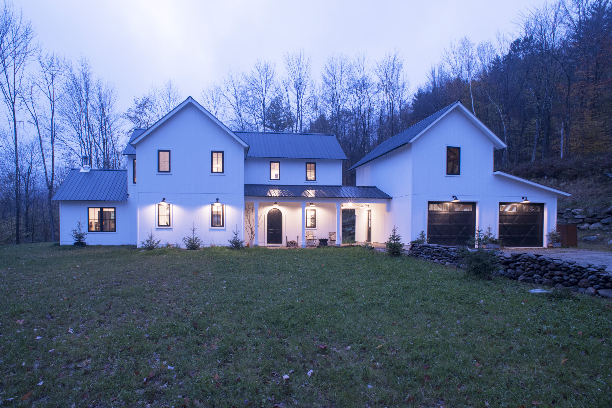 An exterior picture of the Modern New England Farmhouse, designed and built by Geobarns, with white wood siding, black metal roof, detached two-car garage, breezeway, and cupola.