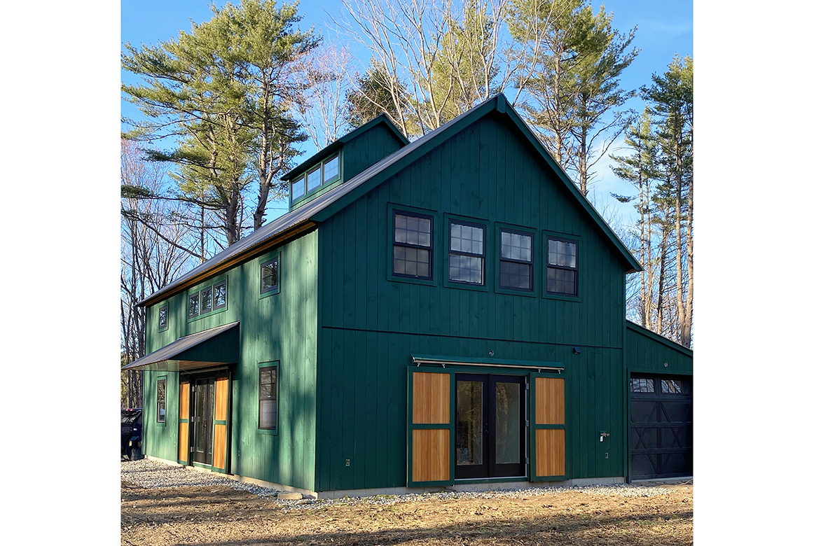 An exterior picture of the Sciobol Barn Retreat, design and built by Geobarns, with jade green wood siding, black metal roof, rolling barn door exterior shutters, and a cupola.