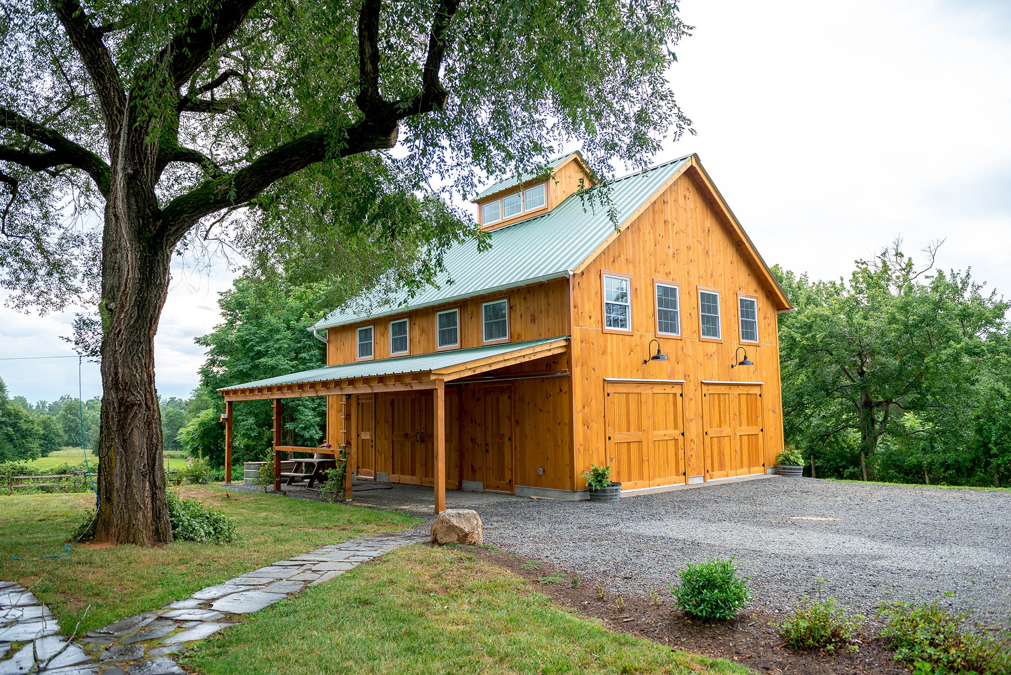 An exterior picture of the Virginia Homestead Barn, design and built by Geobarns, with honey aok stained wood siding, green metal roof, hand-built garage and rolling barn doors, and a monitor cupola.