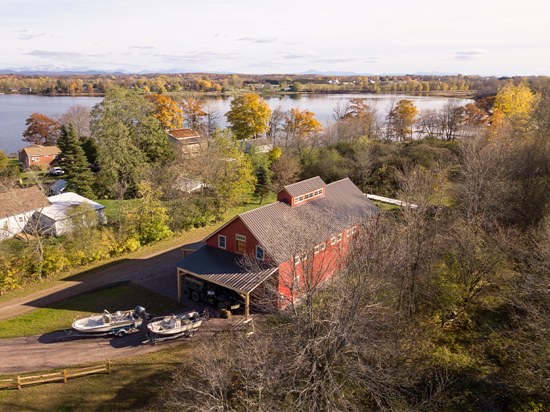 An aerial picture of the Lake Champlain Boat Barn, designed and built by Geobarns, with barn red wood siding, burnished bronze metal roof, lots of room to store and repair boats, and shed awnings for outdoor vehicle storage.