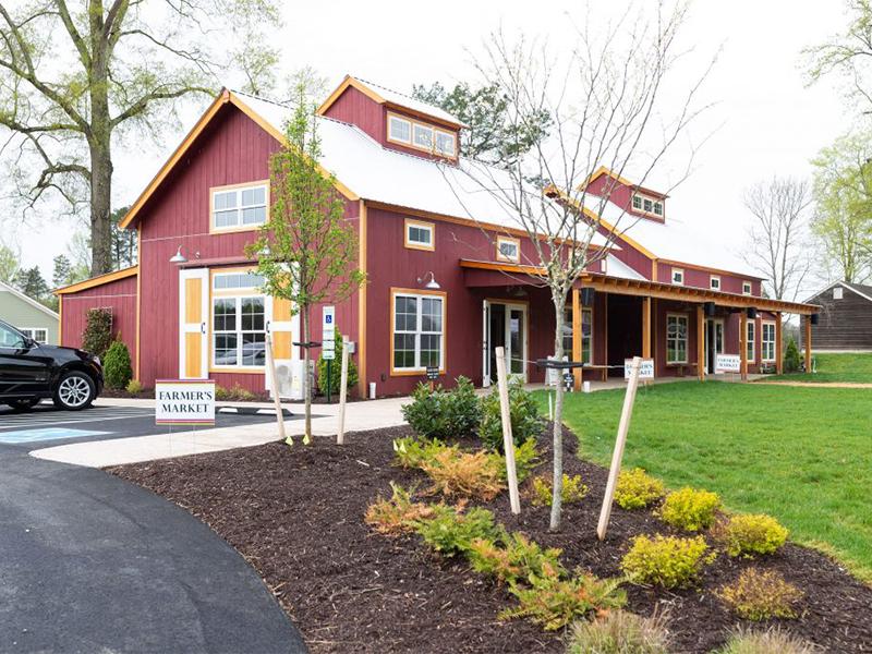 An exterior picture of the sales office and community center of Chickahominy Falls Farm Stand, designed and built by Geobarns, in barn red siding with a silver metal roof, cupolas, and a verandah, next to the community garden.