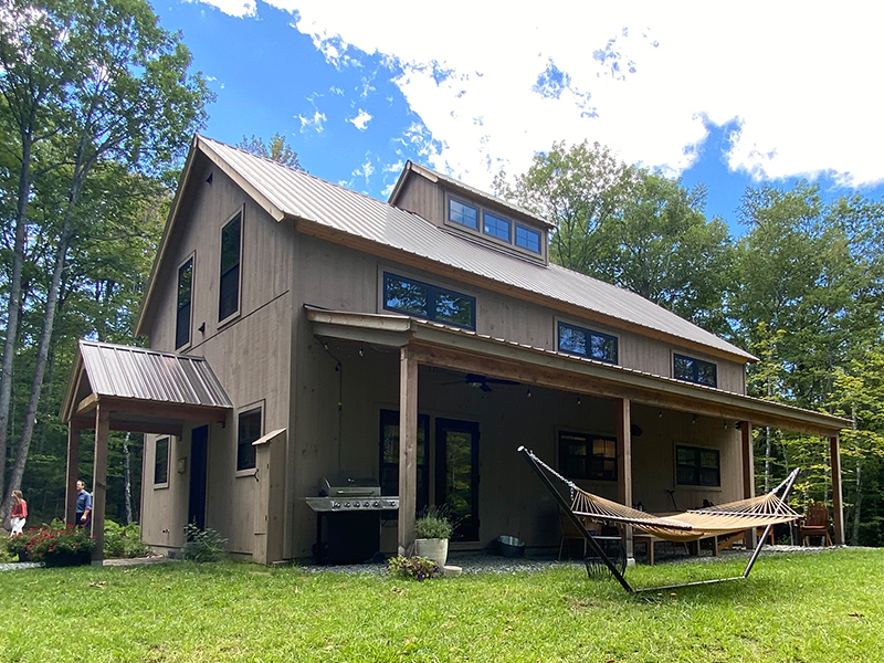 An exterior picture of the Forest Glade Retreat home, designed and built by Geobarns, with mocha wood siding, burnished bronze metal roof, front porch, side porch, and cupola.