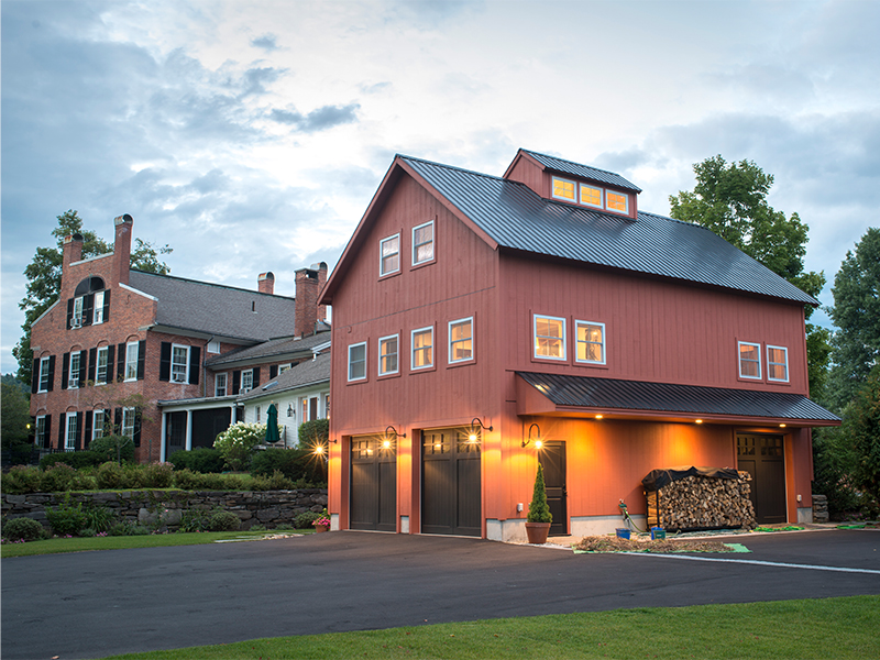 An exterior picture of the Historic Carriage House addition, designed and built by Geobarns, for an historic home in a strict historic district, with barn red siding, black metal roof, and room for cars, recreation space, and home office.