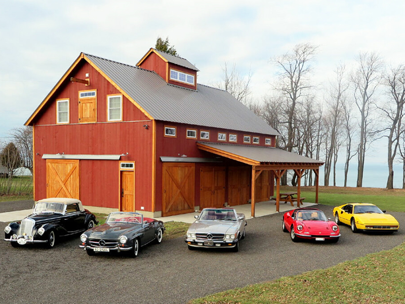 A picture of the Lake Erie Auto Barn, designed and built by Geobarns, with barn red wood siding, gray metal roof, Douglas fir trim, and five sports cars arrayed in front.
