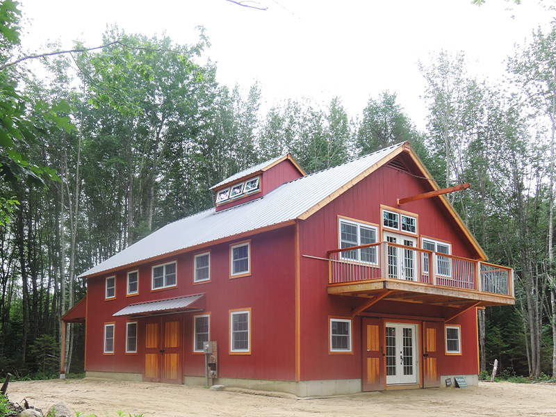 An exterior view of the Maine Coast Barn, designed and built by Geobarns, with barn red wood siding, gray metal roof, wide deack, cupola, and hand-built rolling barn door shutters.