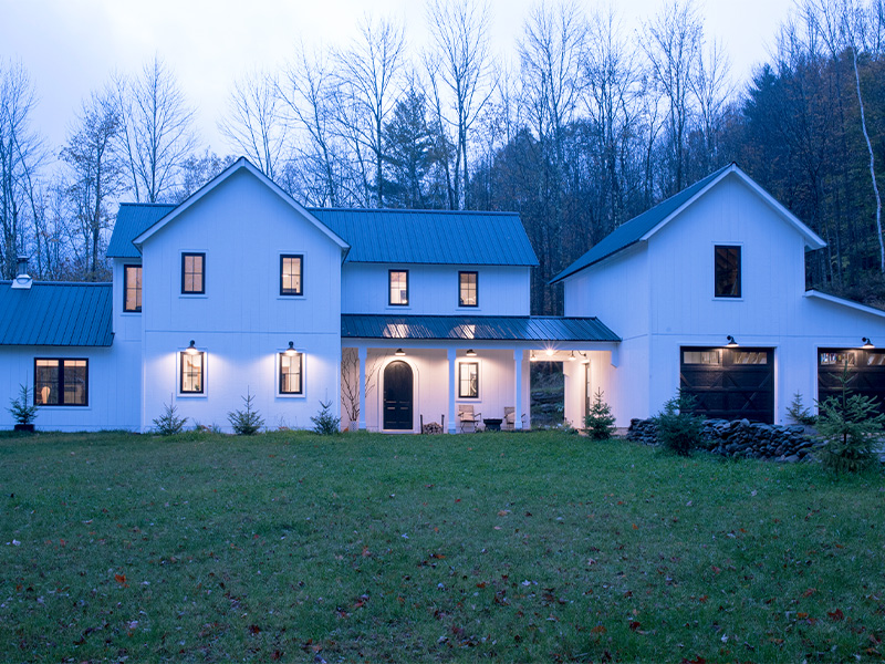 An exterior picture of the Modern New England Farmhouse, designed and built by Geobarns, with white wood siding, black metal roof, detached two-car garage, breezeway, and cupola.