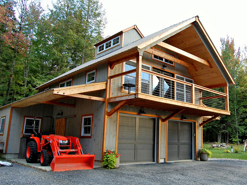 An exterior picture of the Mountain Carriage House, designed and built by Geobarns, with gray wood siding, cantilevered front porch, and side awning to park a Kubota tractor.