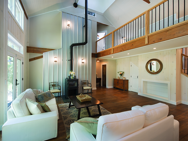 An interior picture of the living room of the Mountaintop Family Home, designed and built by Geobarns, with vaulted ceilings, exposed timber framing, metal firebreak behind a wood stove, mezzanine catwalk, and cupola high above.
