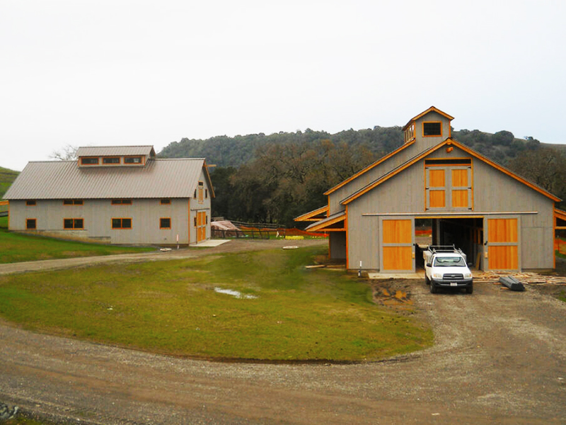 A picture of two Geobarns designed and built to store vehicles and equipment for a farm in California, with gray wood siding and hand-built barn doors with Douglas fir trim.