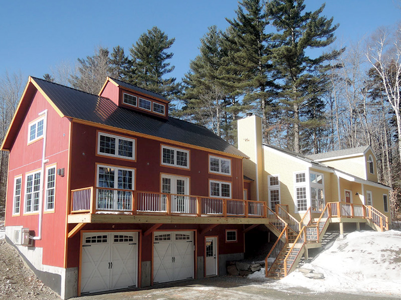 An exterior picture of a garage and office space addition, designed and built by Geobarns, with barn red wood siding, burnished bronze metal roof, hand-built garage doors, a wide porch, and a cupola.