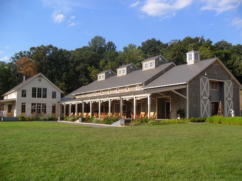 An exterior picture of Pippin Hill Farm and WInery in Virginia, designed and built by Geobarns, showing a wedding hall in gray wood siding, rolling barn door shutters, cupolas, and an attached reception hall designed to look like a Virginia farmhouse.