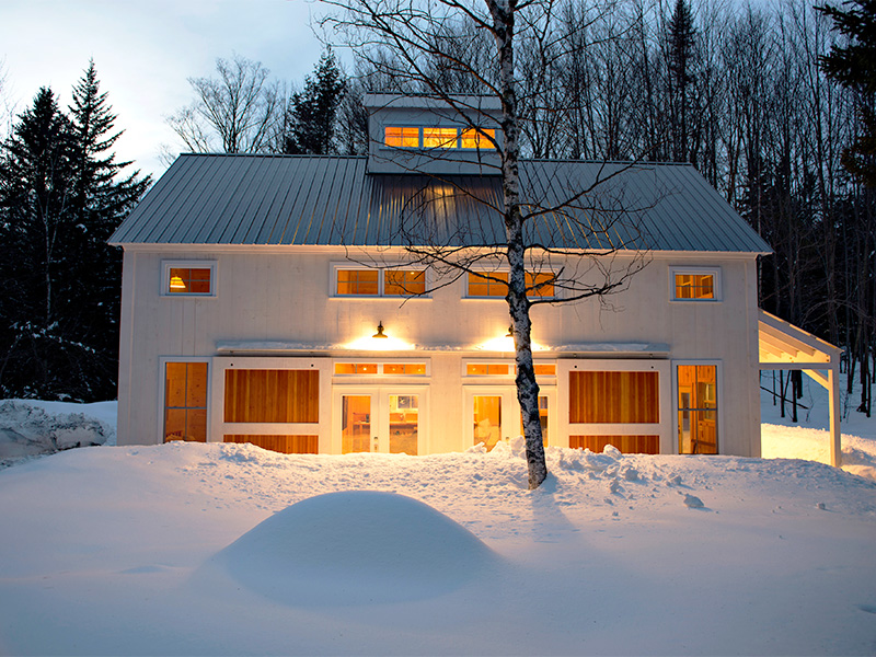 An exterior picture of the Rustic Ski Chalet, deigned and built by Geobarns, with white siding, grey metal roof, and hand-built rolling barn door shutters