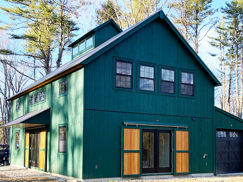 An exterior picture of the Sciobol Barn Retreat, design and built by Geobarns, with jade green wood siding, black metal roof, rolling barn door exterior shutters, and a cupola.