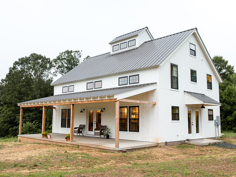 An exterior picture of the Shenandoah Modern Farmhouse, designed and built by Geobarns, with white wood siding, gray metal roof, wide front porch, Douglas fir trim, french door entry, and cupola.
