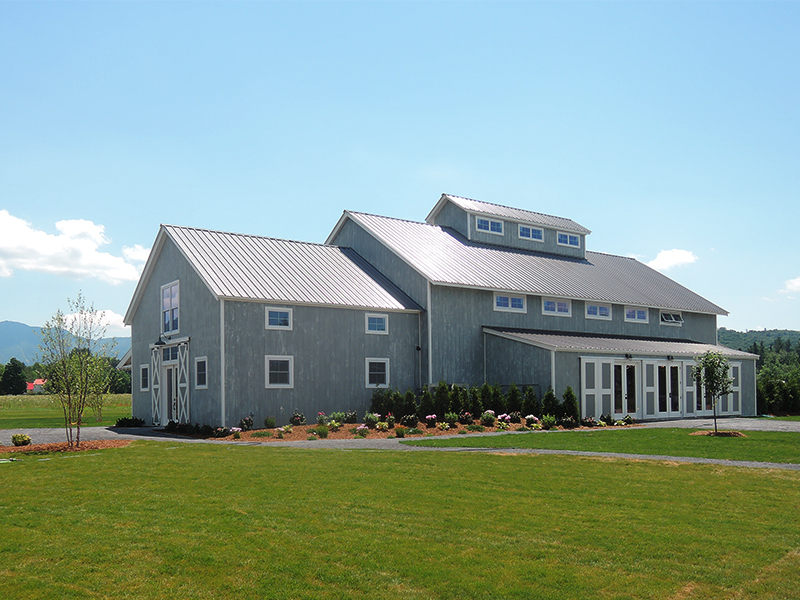 A picture of the Barn at Smuggler's Notch in Stowe, Vermont, a wedding and event barn designed and built by Geobarns, with gray stained wood siding, gray metal roof, cupola, rolling barn doors, event lawn, and Vermont mountains in the background.