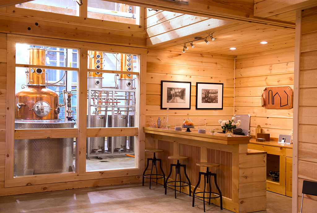 An interior picture of The SILO Distillery tasting room, designed and built by Geobarns, showing shiplap interior wall finishes, a hand-built tasting bar, and windows showing the distillery equipment.