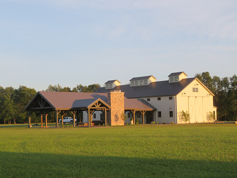 An exterior picture of the Tuck'd Inn Farm wedding barn, designed and built by Geobarns, with whitewashed wood siding, burnished bronze metal roof, three cupolas, and an outdoor timber frame pavilion.