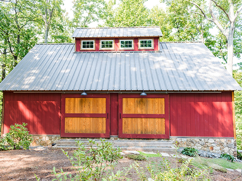 An exteriro picture of the Virginia Car and Shop barn designed and built by Geobarns, with barn red wood siding, gray metal roof, monitor cupola, hand-built rolling barn doors with Douglas fir trim, in a conservation community.