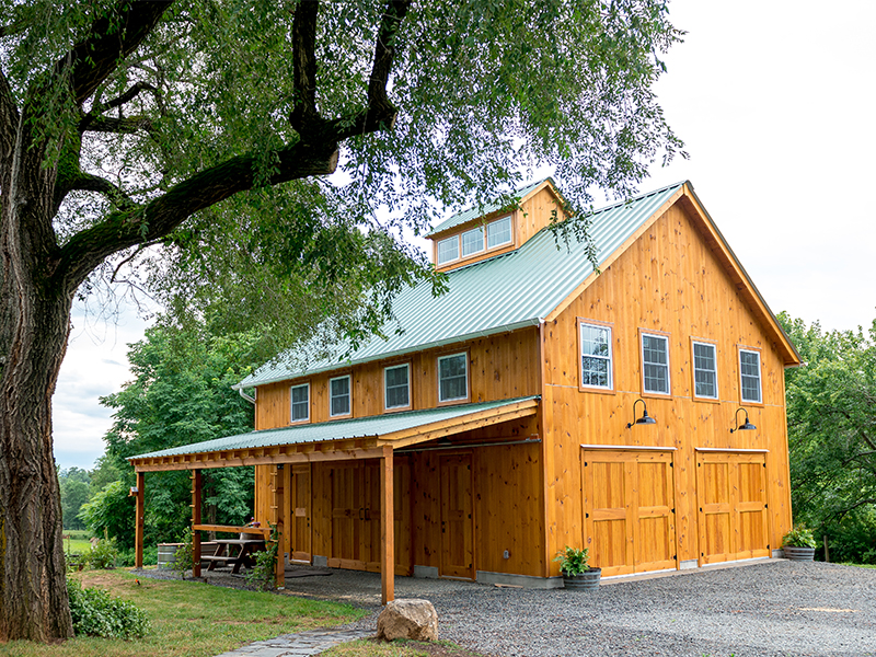 An exterior picture of the Virginia Homestead Barn, design and built by Geobarns, with honey aok stained wood siding, green metal roof, hand-built garage and rolling barn doors, and a monitor cupola.