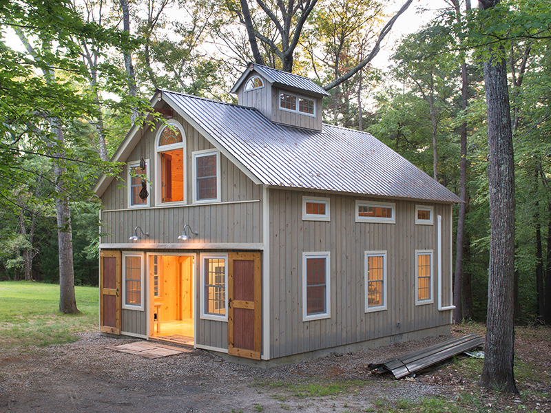 An exterior picture of the Woodworking Temple, design and built by Geobarns, with washed gray wood siding, silver metal roof, rolling barn doors, hayloft door, and cupola.