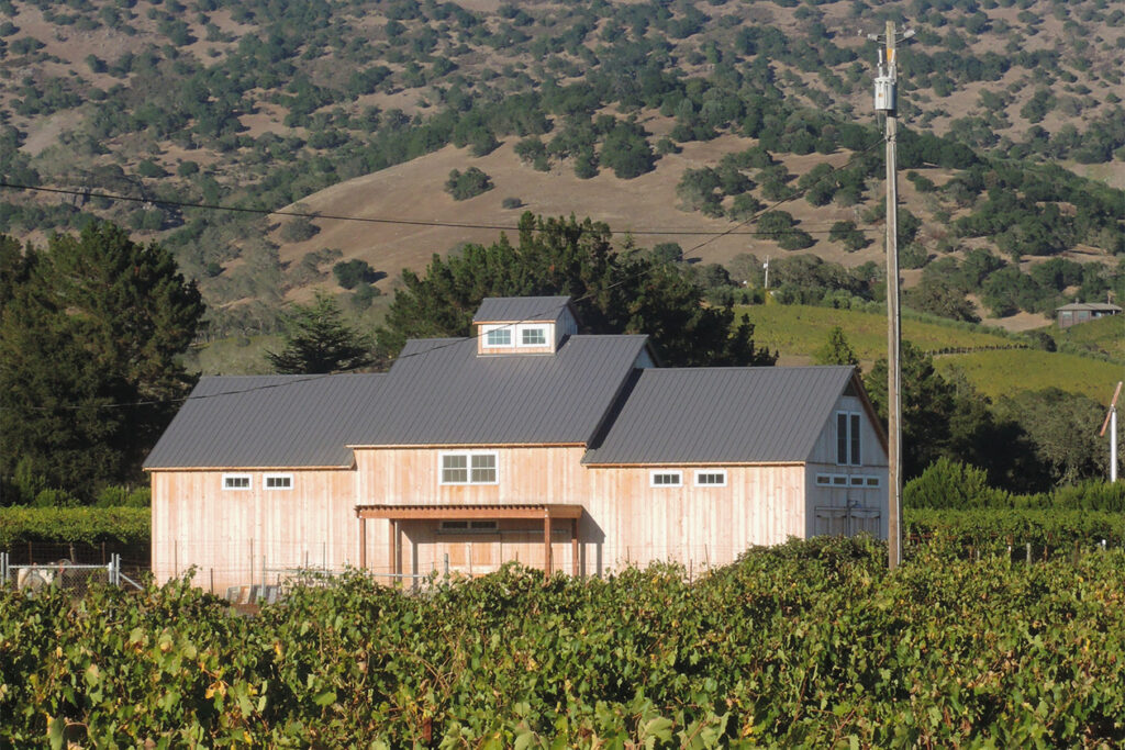 An exterior picture of the Napa Vineyard Barn, designed and built by Geobarns, with natural wood siding and a gray metal roof, in the middle of a vineyard