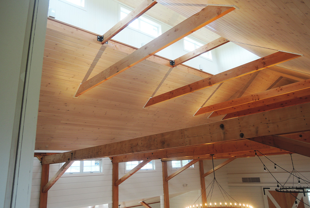 A picture of the the ceiling of a Wedding Barn in Vermont with timber frames, wood ceiling, and sunlight streaming through the cupola