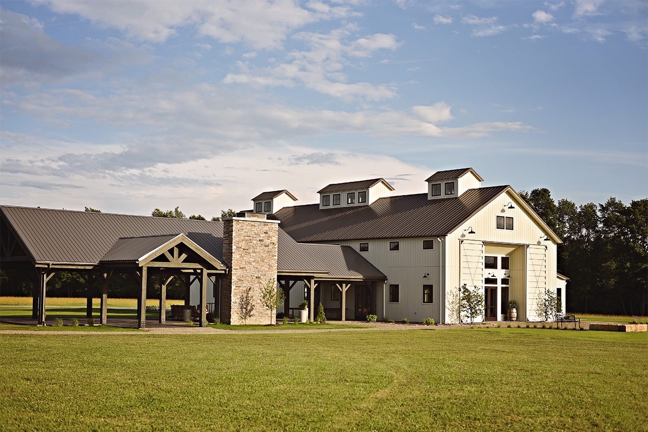 An exterior picture of the Tuck'd Inn Farm wedding barn, designed and built by Geobarns, with whitewashed wood siding, burnished bronze metal roof, three cupolas, and an outdoor timber frame pavilion.