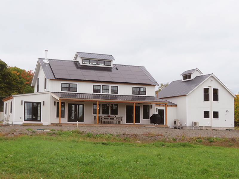 An exterior picture of the Catskills Modern Farmhouse, designed and built by Geobarns, with white wood siding, black metal roof, wide veranda, solar panels, and cupola.